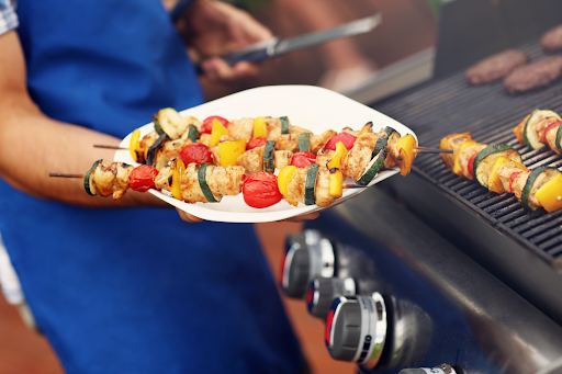 A person holding a plate of grilled kebabs next to a gas barbecue.
